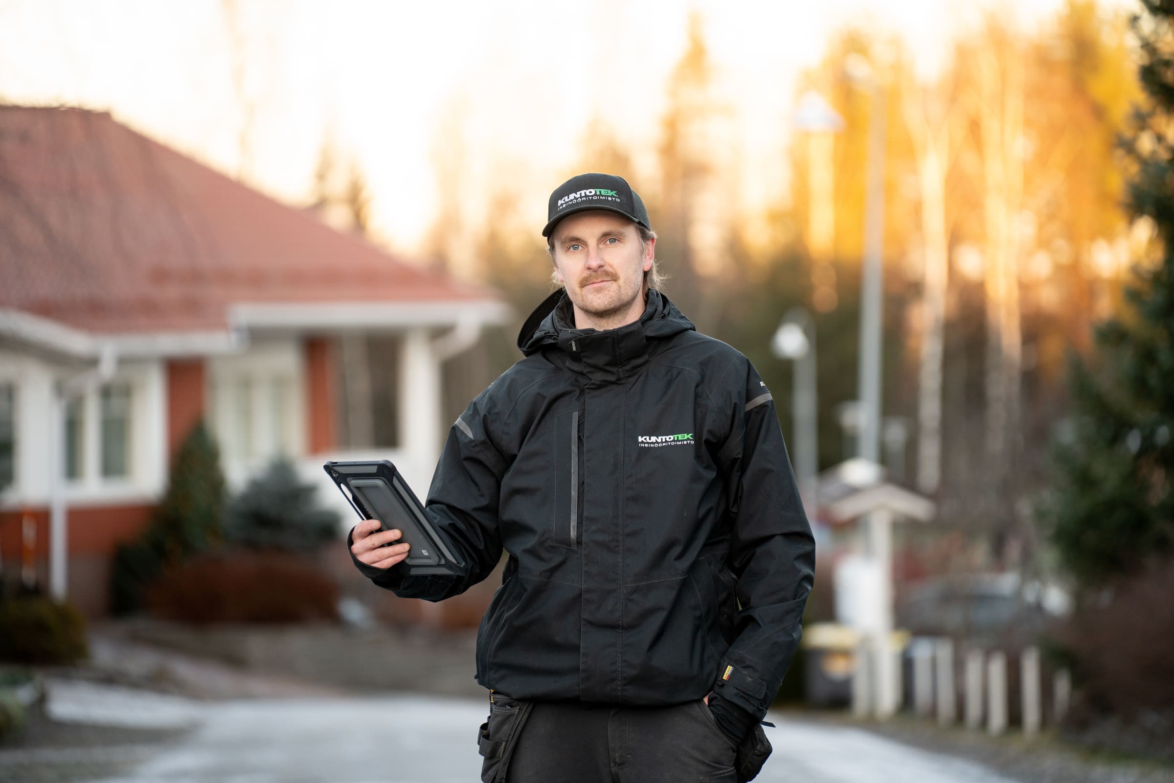Kuntotek Oy:n kuntotarkastaja valmiina tarkastukseen omakotitaloalueella. Asiantuntijalla on käytössään digitaaliset raportointityökalut ja ammattilaisvarustus laadukkaan kuntotarkastuksen varmistamiseksi.