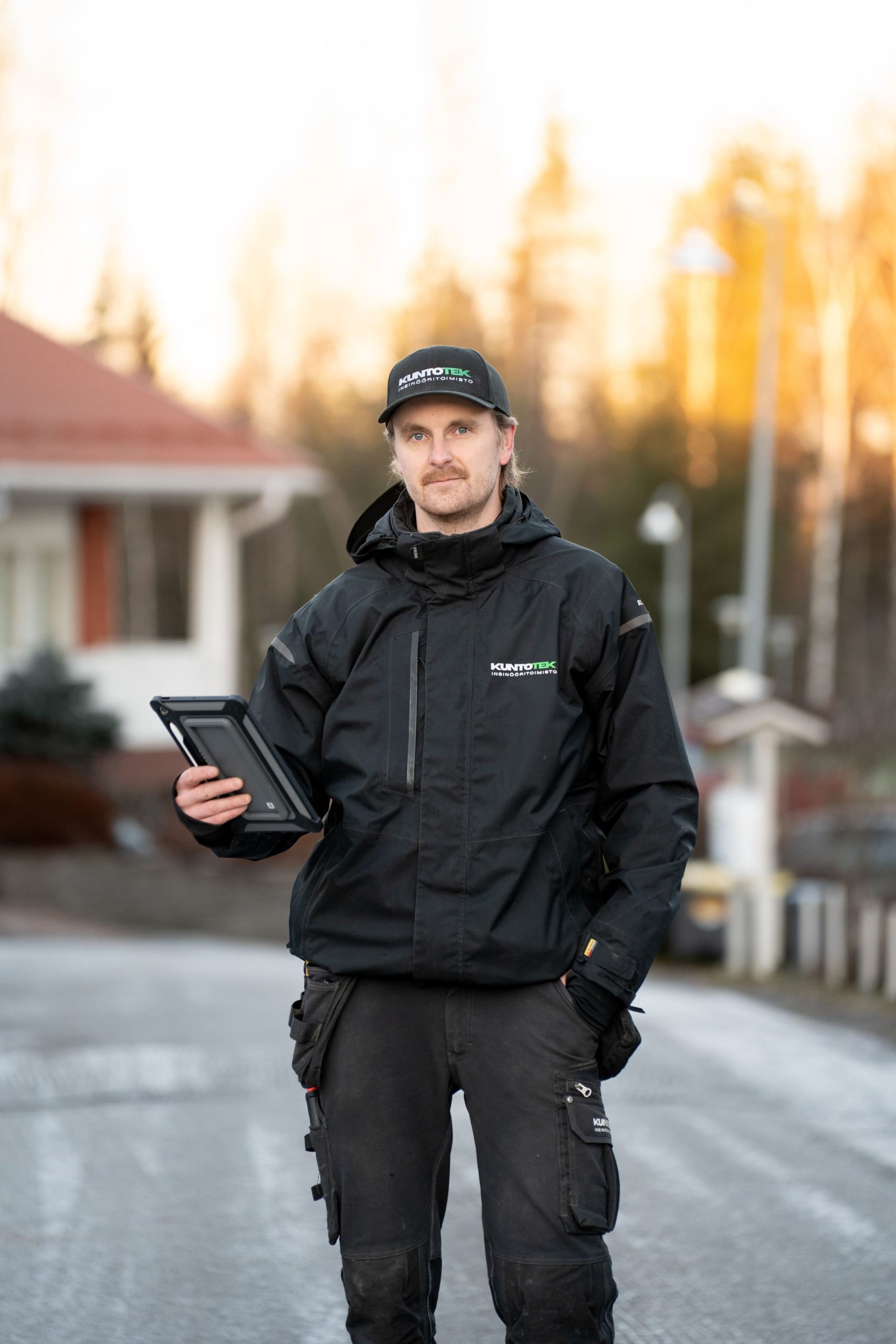 Kuntotek Oy:n kuntotarkastaja valmiina tarkastukseen omakotitaloalueella. Asiantuntijalla on käytössään digitaaliset raportointityökalut ja ammattilaisvarustus laadukkaan kuntotarkastuksen varmistamiseksi.